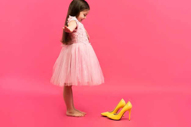 Lustige Kindermädchen Fashionista im Kleid, das die gelben Schuhe der großen Mutter auf farbigem Hintergrund anziehen wird.