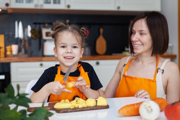 Lustige kleine Tochter und schöne Mutter in der orangefarbenen Schürze kochen, schneiden, hacken Gemüse, lächeln.