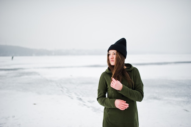 Lustige Mädchenabnutzung auf langem grünem Sweatshirt, Jeans und schwarzem Headwear, in gefrorenem See am Wintertag.