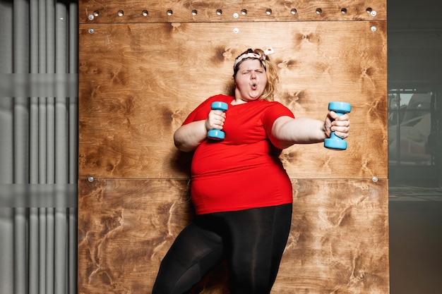 Lustige übergewichtige Frau in Sportkleidung und mit einem Verband auf dem Kopf steht mit Hanteln auf dem Hintergrund einer Holzwand im Fitnessstudio