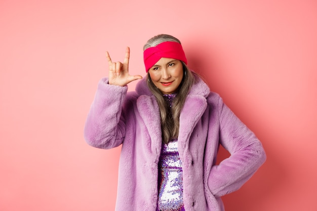 Lustige und coole asiatische Seniorin, die Rock'n'Roll-Geste zeigt, frech aussieht und über rosafarbenem Hintergrund steht.