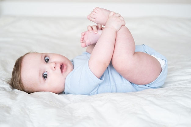 Lustiger 6 Monate kleiner Junge, der mit Beinen spielt und zu Hause auf weißem Bettzeug auf dem Bett liegt.