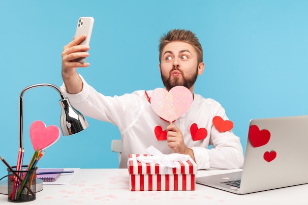 Lustiger bärtiger Büroangestellter, der Selfie macht, Lippen in Kuss drückt und rosa Herz aus Papier hält, am Arbeitsplatz sitzt, alle mit herzförmigen Aufklebern bedeckt. Studioaufnahme im Innenbereich isoliert auf blauem Hintergrund