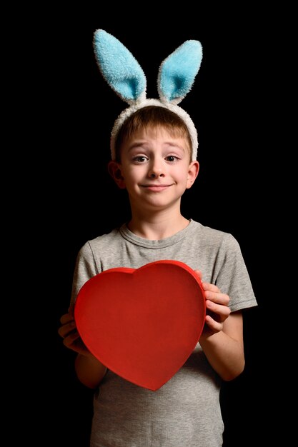 Lustiger blonder Junge in den Hasenohren hält roten Kasten der Herzform auf Schwarzem