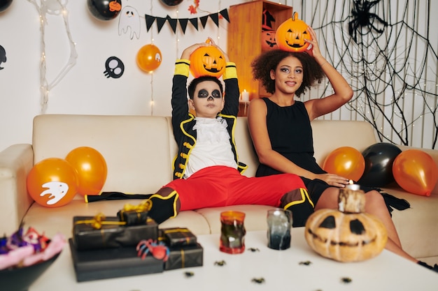 Lustiger Bruder und Schwester mit Halloween-Make-up sitzen auf dem Sofa in einem dekorierten Raum und posieren mit Jack-o-Laternen auf dem Kopf