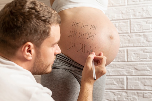 Lustiger Ehemann, der Tage auf Bauch der schwangeren Frau zählt