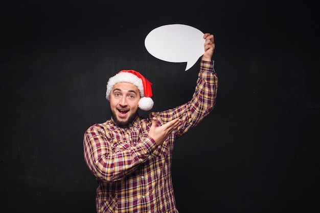 Foto lustiger ernster mann in der roten weihnachtsmann-weihnachtsmütze hält leeren weißen karton als leer oder verspottet mit kopienraum für text. schwarzer hintergrund