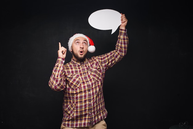 Lustiger ernster Mann in der roten Weihnachtsmann-Weihnachtsmütze hält leeren weißen Karton als leer oder verspottet mit Kopienraum für Text. Schwarzer Hintergrund