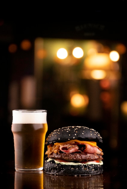 Lustiger Halloween-Themen-Burger, knuspriger schwarzer Brötchenspeck und Zwiebelscheiben mit Sesam auf Holztisch, serviert mit Craft Beer Half Pint mit verschwommenem Pub im Hintergrundporträt