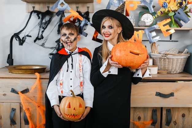 Lustiger Junge und Mädchen, die Halloween-Kostüme tragen, die Kürbisse auf Halloween-Landschaftshintergrund halten. Hochwertiges Foto