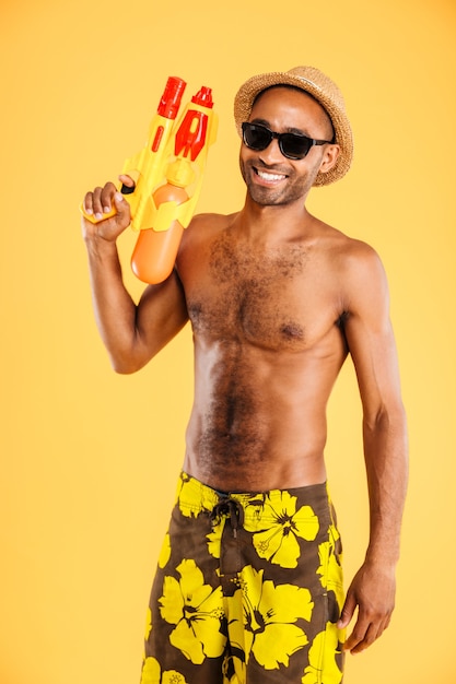 Lustiger junger Afro-Mann in der Badebekleidung, die Wasserpistole über orange Wand hält