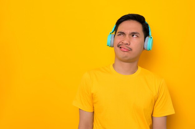 Lustiger junger asiatischer Mann in lässigem T-Shirt, der Musik in Kopfhörern hört und auf den Kopierbereich schaut, der auf gelbem Hintergrund isoliert ist Menschen Emotionen Lifestyle-Konzept