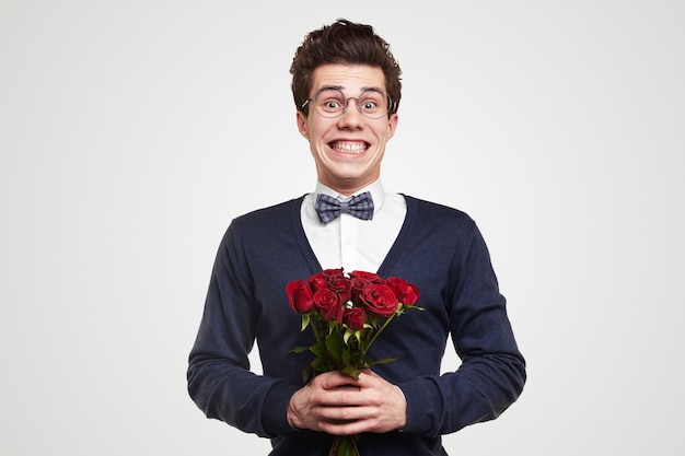 Lustiger junger romantischer Mann in elegantem Anzug und nerdiger Brille mit Strauß roter Rosen, der in die Kamera schaut und hell lächelt, während er zum Valentinstag gratuliert