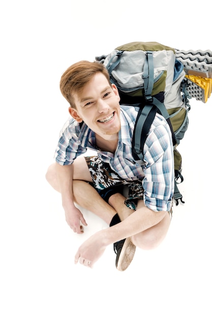 Lustiger junger Tourist sitzt mit Rucksack auf weißem Hintergrund Reisender bereitet sich auf Wanderung vor