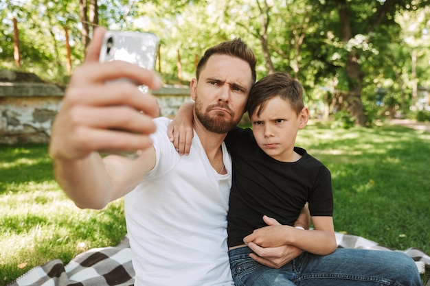 Lustiger junger Vater, der mit seinem kleinen Sohn sitzt, macht Selfie per Handy.