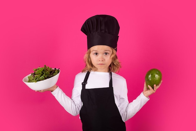 Lustiger Kinderkoch mit Avocado und Gemüse, der Kinder kocht. Koch-Kind-Junge in Form eines Kochs. Kinder-Junge mit Schürze und Kochmütze bereitet in der Küche eine gesunde Mahlzeit zu. Kochvorgang