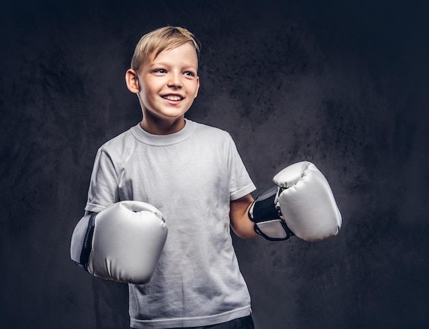 Lustiger kleiner Boxer mit blonden Haaren, gekleidet in ein weißes T-Shirt mit Boxhandschuhen, die in einem Studio posieren. Getrennt auf einem dunklen strukturierten Hintergrund.