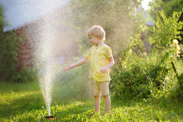 Lustiger kleiner junge, der mit gartenberieselungsanlage im sonnigen hinterhof spielt