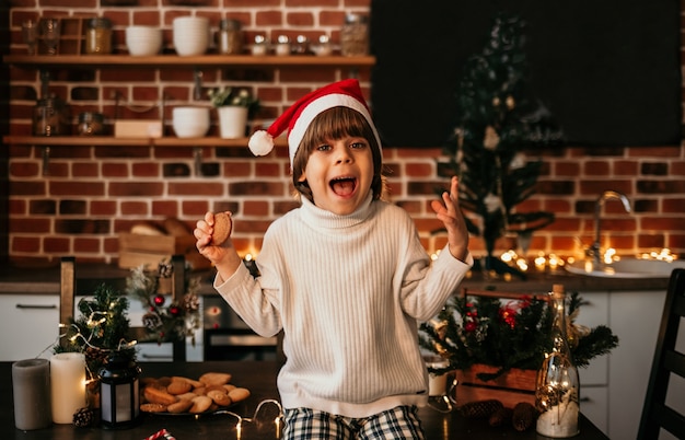 Lustiger kleiner Junge sitzt auf dem Küchentisch in einem weißen Pullover und einer roten Weihnachtsmütze mit Keksen in der Hand