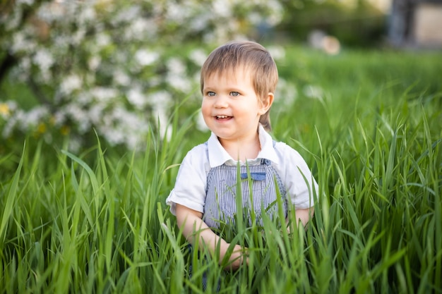 Lustiger lächelnder Junge in denimblauen Overalls und hellblauen Augen. Es ist lustig, sich im hohen grünen Gras in einem warmen Frühlingsgarten vor dem Hintergrund blühender Bäume zu verstecken.