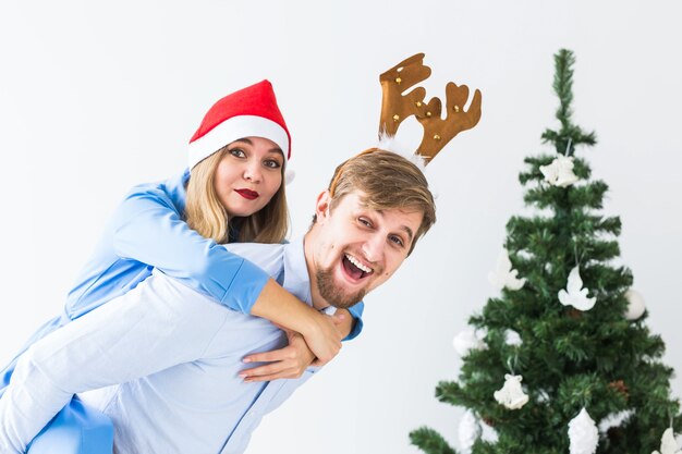 Lustiger Mann, der seiner Frau Huckepack gibt, während sie zu Hause Weihnachtsmützen für die Weihnachtsfeiertage tragen.