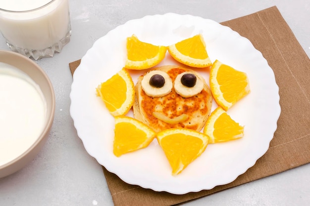 Lustiger Sonnengesichtsform-Snack aus Pfannkuchenorange auf dem Teller Süßes Dessert für süße Kinder, gesundes Frühstück, Mittagessen, Lebensmittelkunst mit Milch und Sauerrahm auf grauem Betonhintergrund, Ansicht von oben