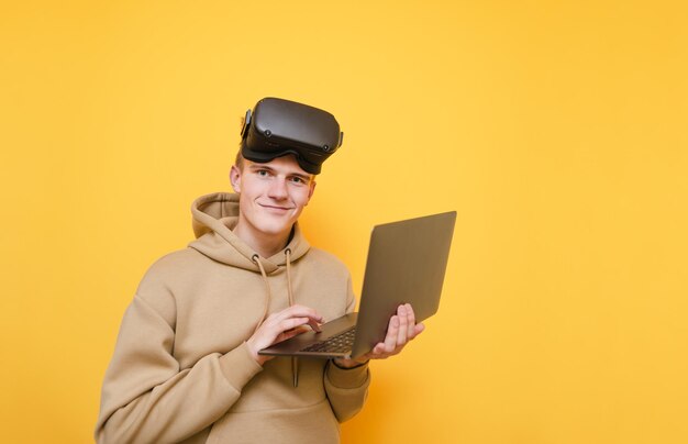 Lustiger Teenager in legerer Kleidung steht auf gelbem Hintergrund mit einem Laptop in den Händen mit VR-Brille auf dem Kopf schaut mit einem Lächeln in die Kamera Isoliertes VR-Virtual-Reality-Konzept