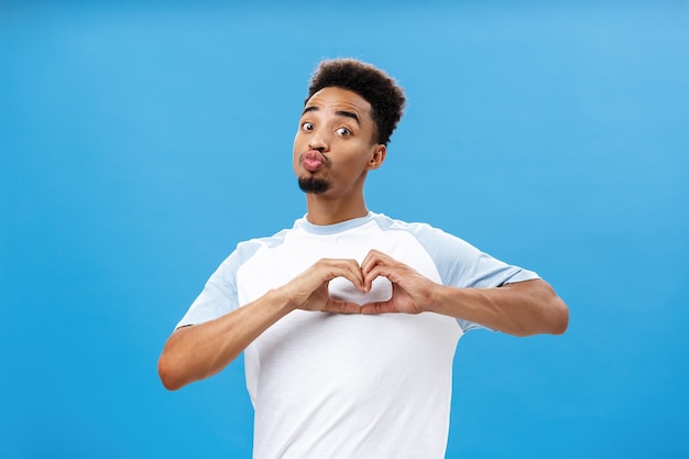 Lustiger und süßer dunkelhäutiger junger männlicher Student im T-Shirt, der Lippen in Mwah faltet oder Kuss mit Herz g ...