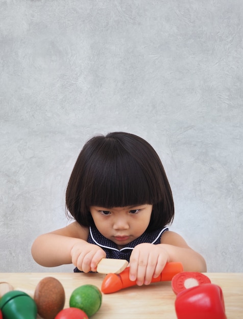 Lustiges asiatisches Kindermädchen, das mit hölzernem kochendem Spielzeug, kleiner Chef zubereitet Lebensmittel auf Küchenarbeitsplatte spielt.