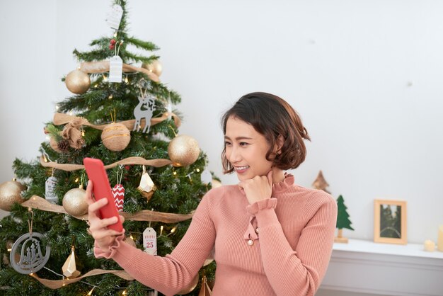 Lustiges asiatisches Mädchen, das zu Hause in der Nähe des Weihnachtsbaums Selfie-Bilder auf der Smartphone-Kamera macht