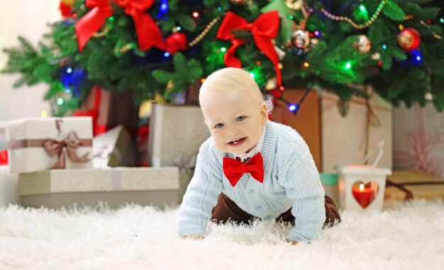 Lustiges Baby auf weichem Teppich und Weihnachtsbaum auf der Oberfläche