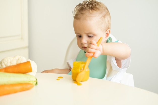 Lustiges Baby im Lätzchen, das Gemüsepüree mit Löffel isst, der im Kinderstuhl sitzt, lernt zu essen