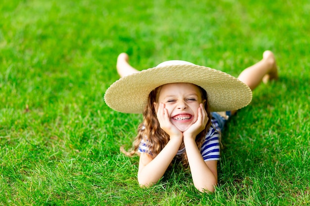 Lustiges Baby im Sommer auf dem Rasen mit einem großen Strohhut auf dem grünen Gras, das Spaß hat und sich freut, Platz für Text