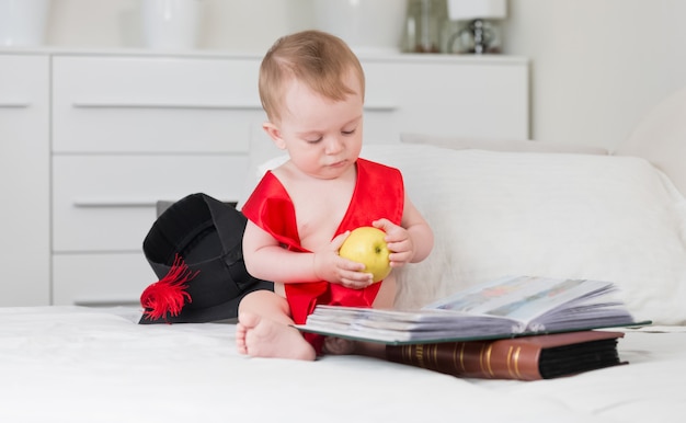 Lustiges Baby in Abschlusskappe und Band, das Apfel hält und großes Buch liest