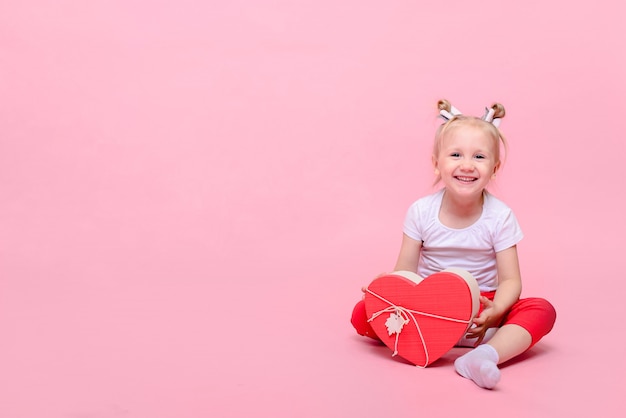 Lustiges Baby in einem weißen T-Shirt und in der roten Hose mit einer herzförmigen Schachtel auf einem rosa Hintergrund.