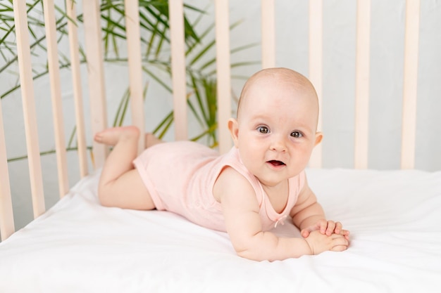 Lustiges Babymädchen in einer Krippe in einem rosafarbenen Bodysuit für sechs Monate auf einem weißen Baumwollbett liegt auf dem Bauch und lächelt morgens aufgewacht oder geht ins Bett