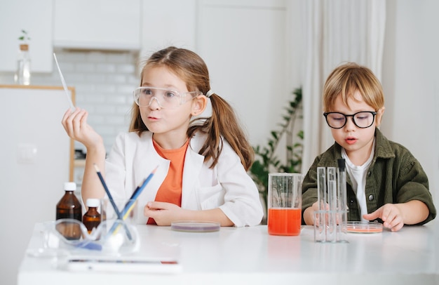 Lustiges emotionales kleines Mädchen im weißen Gewand, das eine Pipette hält, neben ihrem Jungen wählt ein Gericht mit roter chemischer Flüssigkeit aus. Machen Sie ein Home-Science-Projekt hinter dem Tisch.