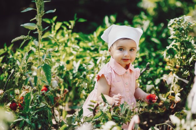 Lustiges, entzückendes Baby mit weißem Kopftuch, das Tomate im Garten isst und in die Kamera lächelt