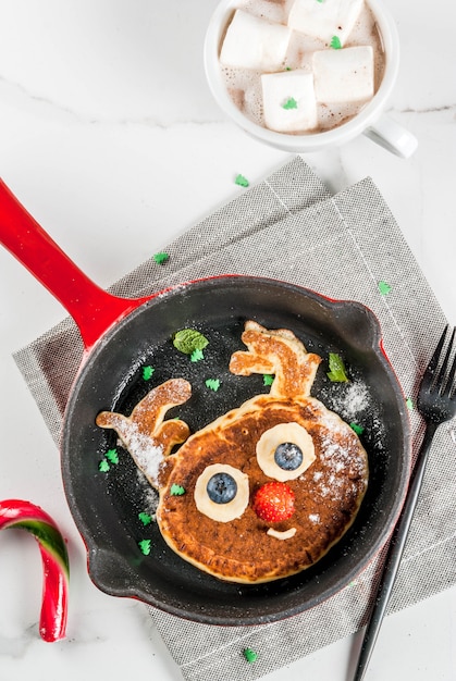 Lustiges Essen zu Weihnachten. Kinderfrühstückspfannkuchen verziert wie Ren, mit heißer Schokolade mit Eibisch, weiße Tabelle