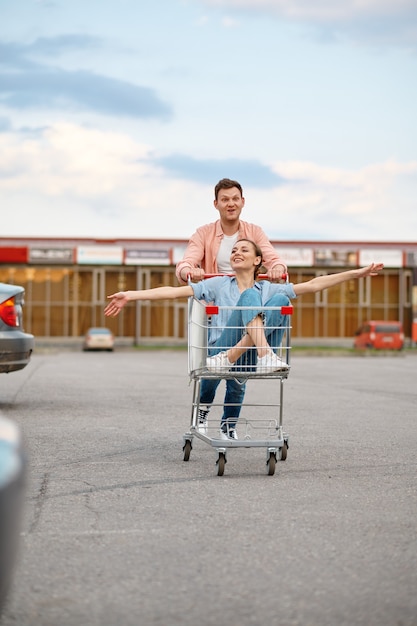 Lustiges Familienpaar fährt im Wagen auf dem Parkplatz des Supermarkts. Glückliche Kunden, die Einkäufe aus dem Einkaufszentrum tragen, Fahrzeuge im Hintergrund, Mann und Frau auf dem Markt