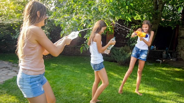 Lustiges Foto einer glücklichen Familie mit Kindern, die am heißen sonnigen Tag mit Wasserpistolen und Gartenschlauch spielen und Wasser spritzen