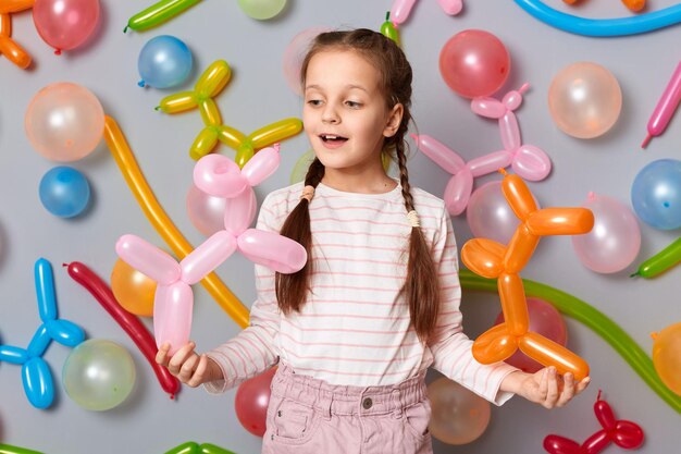 Lustiges Geburtstagskind Spielzeit Kleines Mädchen mit Zöpfen, das an einer grauen Wand steht, die mit bunten Luftballons geschmückt ist, lächelt glücklich und genießt den Urlaub