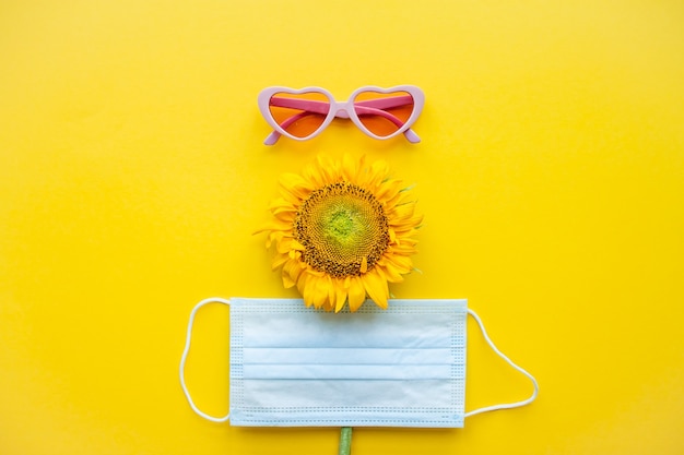 Lustiges Gesicht aus rosafarbener herzförmiger Sonnenbrille, einer medizinischen Schutzmaske und einer Sonnenblume auf einem hellen