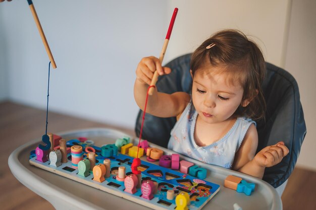 Lustiges glückliches Baby, das auf einem Hochstuhl sitzt und mit pädagogischem Holzspielzeug spielt Konzept des frühen Lernens und der Entwicklung feinmotorischer Fähigkeiten