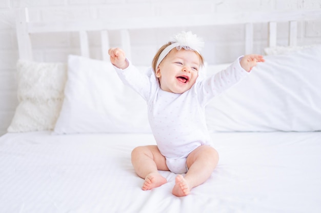 Lustiges, glückliches Mädchen, das in weißen Kleidern auf dem Bett sitzt und ihre Hände hochhält, tanzt oder sich streckt, nachdem sie morgens geschlafen hat, ein kleines Kind auf einem Baumwollbett zu Hause lächelnd