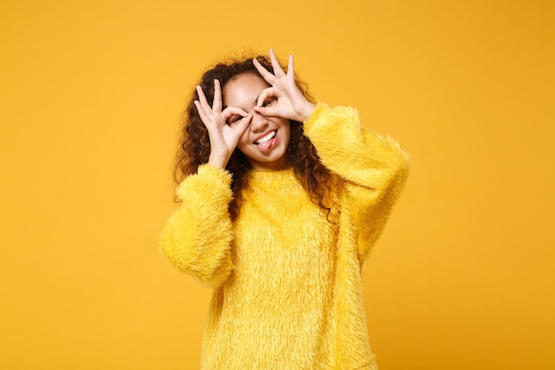 Lustiges junges afrikanisch-amerikanisches Mädchen, das isoliert auf gelb-orangefarbenem Hintergrund posiert. People-Lifestyle-Konzept. Mock-up-Kopienbereich. Zunge zeigen, Hände nahe an Augen halten, Brille oder Fernglas imitieren.
