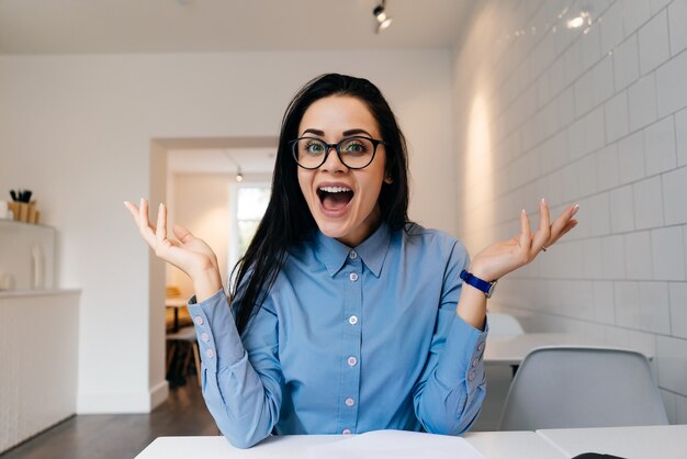 Lustiges junges Mädchen in blauem Hemd und Brille lacht, sitzt im Café