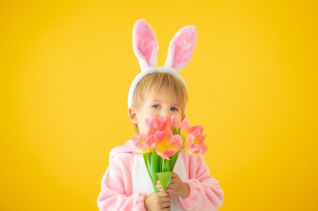 Lustiges Kind, das Osterhasenohren trägt und einen Strauß Tulpen gegen gelbe Wand hält.