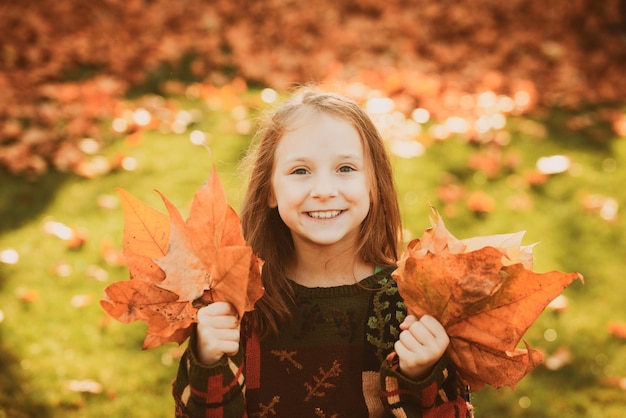 Lustiges Kind draußen im Herbstpark. Entzückendes glückliches Mädchen, das mit gefallenen Blättern im Herbstpark spielt