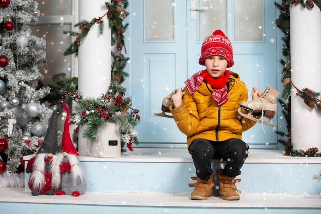 Lustiges Kind hält alte Schlittschuhe und sitzt auf der Veranda eines mit Weihnachtsbaum geschmückten Hauses